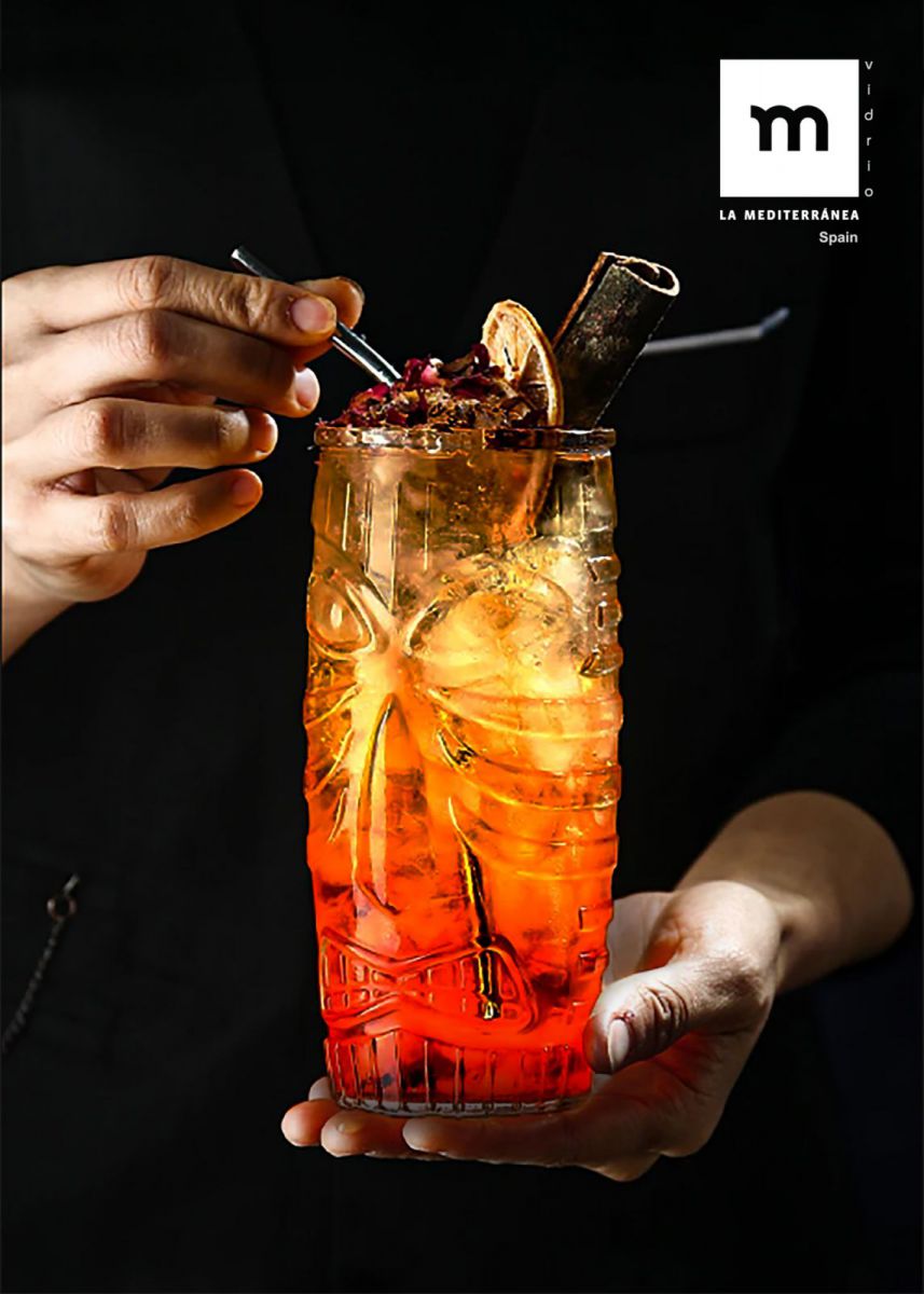 Vibrant orange cocktail in textured Tiki Highball glass garnished with dried fruit berries and cinnamon stick