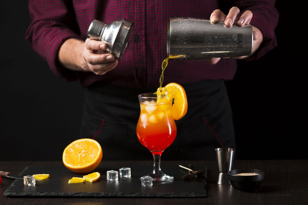 Bartender pouring vibrant orange and red cocktail into glass with ice and orange slice